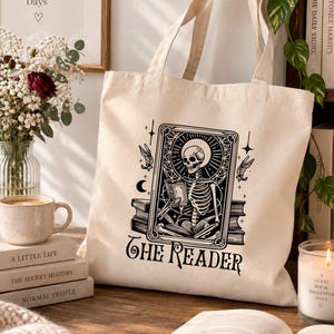 Peut inclure: Un sac fourre-tout en toile couleur crème avec une impression graphique noire représentant un squelette lisant un livre dans un cadre rectangulaire. Le motif inclut des étoiles, un croissant de lune et deux oiseaux, avec le texte THE READER.