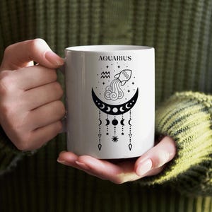 Könnte beinhalten: Weiße Keramiktasse mit dem Wort "AQUARIUS" über einem schwarzen Halbmond-Design, Sternen und einer Wasserkanne-Illustration. Die Tasse wird von einer Person gehalten, die einen grünen Pullover trägt. Das Design umfasst hängende Himmelselemente.