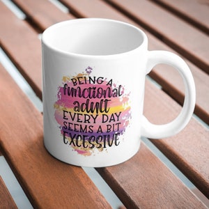 K&ouml;nnte beinhalten: Wei&szlig;er Keramikbecher mit Henkel. Der Becher zeigt den Text "Being a functional adult every day seems a bit excessive" in einem farbenfrohen Aquarelldesign mit rosa, lila, gelben und goldenen Akzenten.