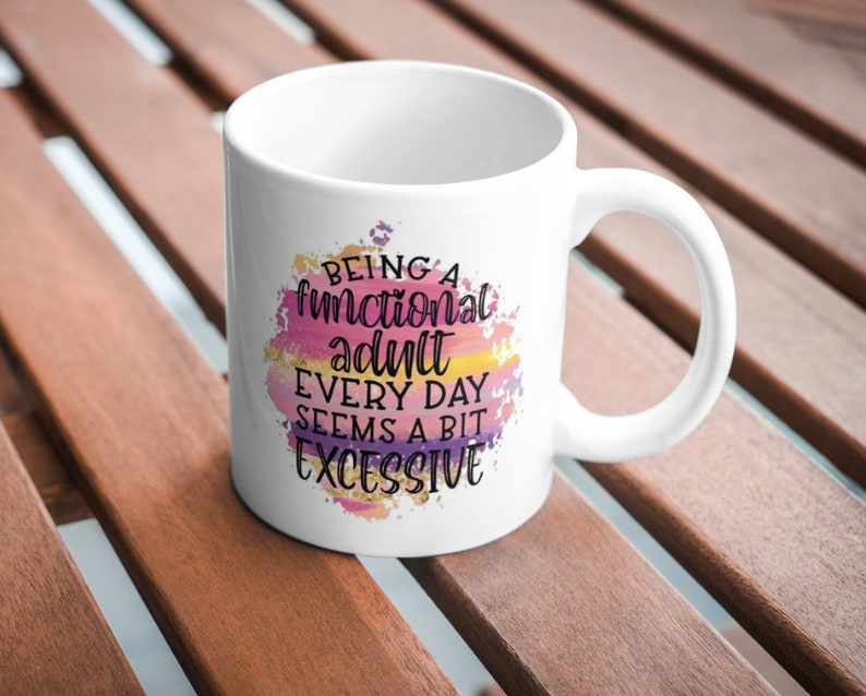 K&ouml;nnte beinhalten: Wei&szlig;er Keramikbecher mit Henkel. Der Becher zeigt den Text "Being a functional adult every day seems a bit excessive" in einem farbenfrohen Aquarelldesign mit rosa, lila, gelben und goldenen Akzenten.