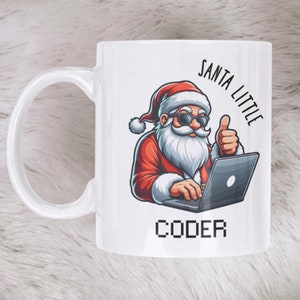 Puede incluir: Taza de cerámica blanca con un diseño de Papá Noel de dibujos animados con traje rojo y gafas de sol, usando un portátil. La taza tiene el texto "SANTA LITTLE CODER". La taza está sobre una superficie blanca y esponjosa.