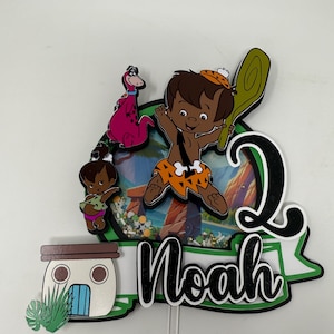 Puede incluir: Un adorno para pastel verde, negro y blanco con un personaje de dibujos animados de los Picapiedra, un dinosaurio, una casa y el nombre "Noah".