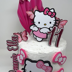 May include: A white frosted cake decorated with Hello Kitty figures, pink bows, and butterfly accents. The cake is adorned with the number 11 and the names "Ella" and "Amerie" in pink and black lettering. A festive birthday cake.