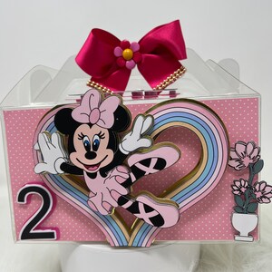 Puede incluir: Una caja de regalo rosa y blanca con una tapa transparente. La caja tiene un diseño de personaje de Minnie Mouse con un corazón de arcoíris y el número 2. La caja tiene un lazo rosa con una flor amarilla en la parte superior.