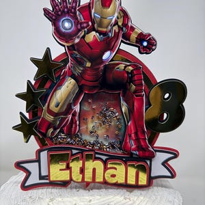 May include: A red and gold Iron Man cake topper with a banner that says "Ethan" and a gold number 8. The topper is on a white cake.