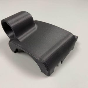 Puede incluir: Una pieza de plástico negro impresa en 3D con una parte superior redondeada y una base plana. La pieza tiene un agujero en la parte superior y una serie de crestas en la parte inferior.