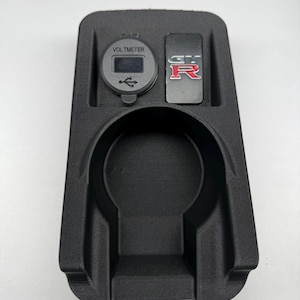 May include: Black automotive console with a voltmeter, USB port, and a GT-R emblem. The console has a cup holder and a rectangular slot for a switch. The voltmeter displays a digital readout.
