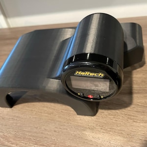 Può includere: Un indicatore digitale Haltech nero montato su una staffa stampata in 3D nera. L'indicatore ha una ghiera nera con un logo Haltech giallo e un display digitale.