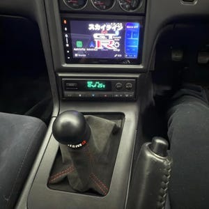 Op de afbeelding: Een zwart lederen auto-interieur met een zwarte en zilveren versnellingspook, een zwarte leren hoes en een digitaal display met de tekst "スカイライン GT-R" in het Japans.