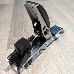 May include: A racing simulator pedal assembly featuring a silver metal frame with black accents. The unit includes a vertical pedal plate, a horizontal spring-loaded tension mechanism with a black housing, and exposed metal bolts and circular cutouts.