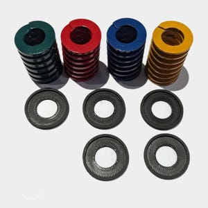 Może przedstawiać: Asortyment sprężyn i podkładek przemysłowych. Sprężyny są w kolorze zielonym, czerwonym, niebieskim i złotym. Podkładki są czarne z okrągłym wzorem i centralnym otworem. Przedmioty są ułożone na białej powierzchni.