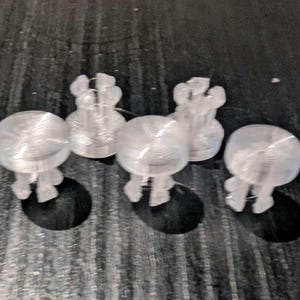 Op de afbeelding: Een set van vijf doorzichtige, doorschijnende kunststof bevestigingsmiddelen, elk met een platte ronde bovenkant en een basis met meerdere tanden voor insteekmontage. Deze kleine, stijve onderdelen hebben een gladde, gelaagde textuur.