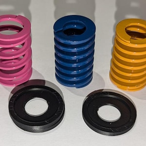 May include: Three metal springs in pink, blue, and yellow colors, and two black washers. The springs are coiled and have a circular shape. The washers are flat and have a hole in the center.