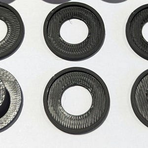 May include: Six black plastic rings with a raised inner ring. The rings have a textured surface and are designed to be used as a spacer or a decorative element.