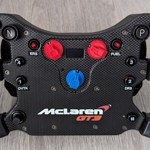 May include: A black McLaren GT3 racing wheel with multiple buttons and dials. Features red and blue controls, and the McLaren logo. The wheel has a carbon fiber texture and is designed for racing simulation.