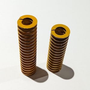 May include: Two yellow-gold compression springs. Each spring is cylindrical, with a series of circular coils. The top of each spring has a split ring design. The springs are set against a white background.