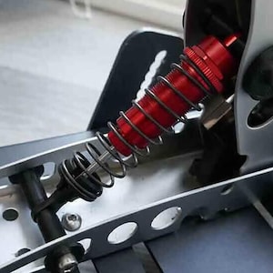 Puede incluir: Un amortiguador rojo y plateado con un resorte helicoidal. El amortiguador está unido a un marco de metal.