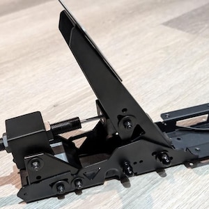 Puede incluir: Un conjunto de pedales de metal negro para simulador de carreras que cuenta con un brazo de pedal vertical, un mecanismo de tensión horizontal con resorte y varilla roscada, y una base de montaje plana con múltiples orificios.