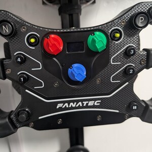 May include: A black Fanatec racing steering wheel with a carbon fibre texture finish. It features multiple buttons labelled with numbers and symbols, including a central display, and three prominent rotary dials in red, green, and blue colours.