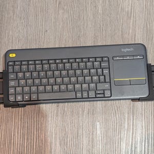 May include: A dark grey Logitech wireless keyboard with a built-in touchpad. The keyboard has a full QWERTY layout with black keys and white lettering. A yellow button is visible in the upper left corner.