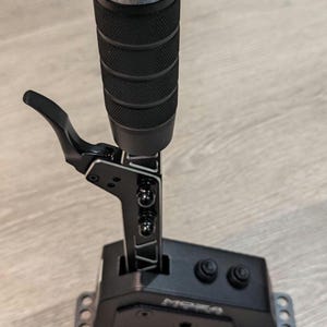 Puede incluir: Palanca de cambios negra con una perilla negra y una palanca plateada. La palanca de cambios tiene una base negra con la palabra "MPSA" impresa en ella. Hay dos botones negros en la base.