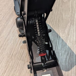 Op de afbeelding: Close-up van een zwart metalen mechanisme met een rood-zwarte veer. Het mechanisme is bevestigd aan een zwarte metalen basis.