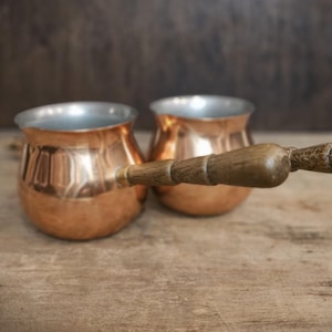 May include: Two copper coffee pots with wooden handles. The pots are sitting on a wooden surface.