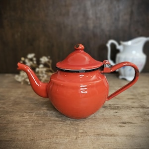 Op de afbeelding: Een klein, rood emaille theepotje met een zwarte rand en een scharnierend deksel. Het theepotje heeft een rond lichaam en een gebogen tuit.