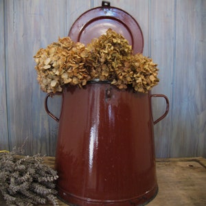 May include: A maroon enamelware pot with a lid, filled with dried hydrangeas. The pot has two side handles and sits on a wooden surface. A bundle of dried lavender is to the left of the pot. The pot is a deep red colour.