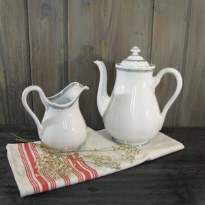 May include: A white porcelain teapot and creamer set with a delicate gray trim. The teapot has a lid and a spout, while the creamer has a handle. Both pieces are sitting on a red and white striped cloth.