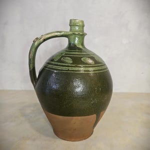 May include: A dark green ceramic jug with a handle and a spout. The jug has a bulbous body and a lighter brown base. Decorative green patterns encircle the top of the jug. The jug is likely an antique or vintage piece.