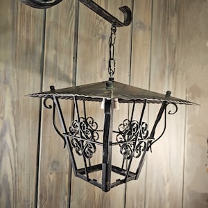May include: A black metal lantern with a square shape and intricate scrollwork details hangs from a chain attached to a metal hook. The lantern has a flat top and a decorative design.