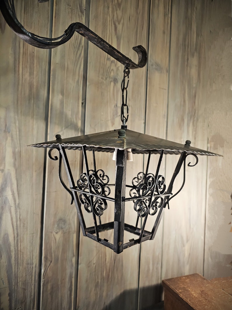 May include: A black metal lantern with a square shape and intricate scrollwork details hangs from a chain attached to a metal hook. The lantern has a flat top and a decorative design.