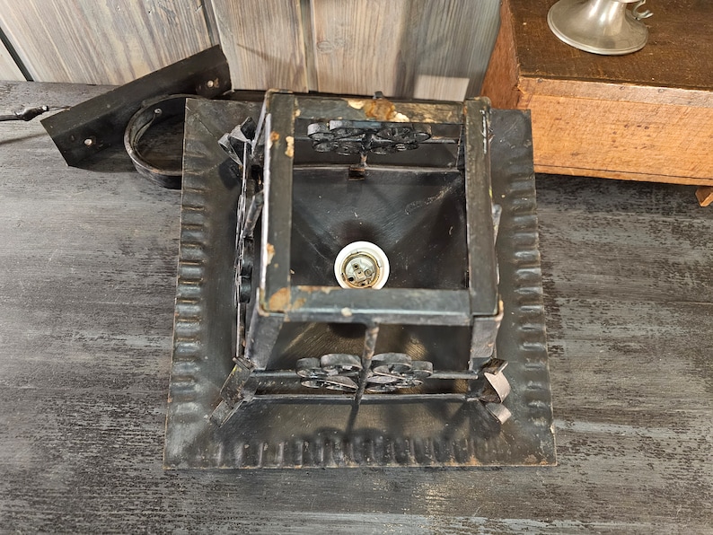 May include: A black metal ceiling light fixture with a square base and a decorative design. The light fixture has a single bulb socket and a clear glass bulb.