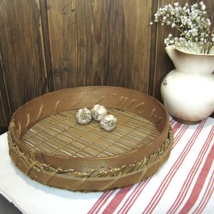 May include: A round, wooden, woven basket with a bamboo insert. The basket has a natural brown color and is sitting on a red and white striped tablecloth. There are four garlic cloves inside the basket.