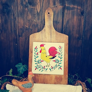Op de afbeelding: Een houten snijplank met een haanontwerp. De haan is geel met een rode kam en rode keellap, en een rode staart. De snijplank heeft een handvat en is versierd met groene bladeren en rode bloemen.