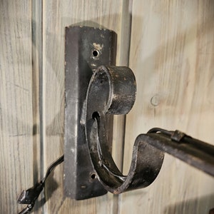 May include: Black metal wall mount bracket with a decorative scroll design. The bracket is attached to a wooden wall.