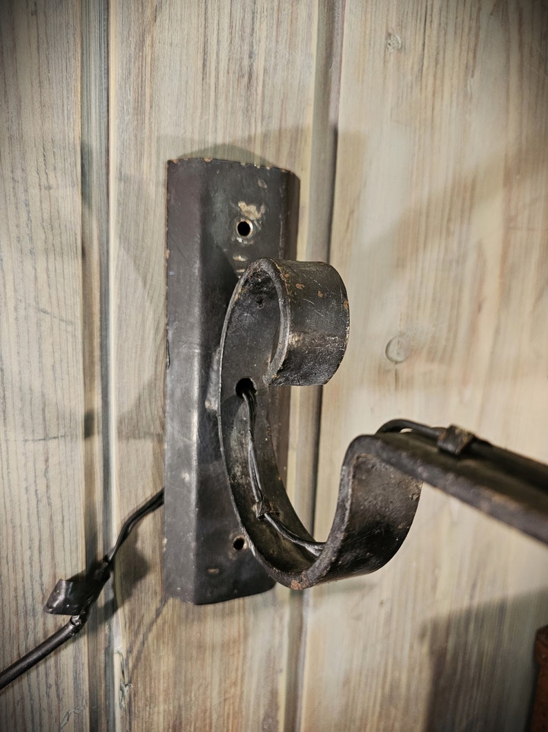 May include: Black metal wall mount bracket with a decorative scroll design. The bracket is attached to a wooden wall.