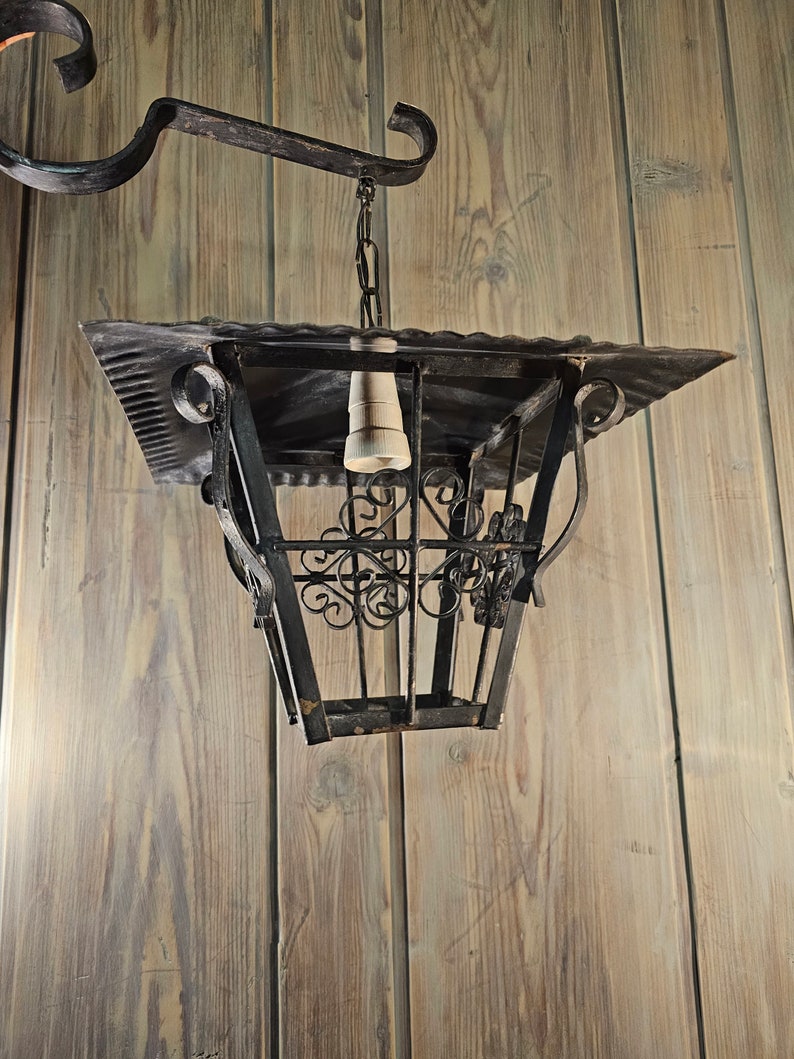 May include: A black metal lantern with a decorative design hangs from a chain attached to a hook on a wooden wall.