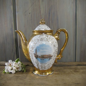 May include: A white porcelain teapot with gold accents and a floral design. The teapot features a painted scene of Venice, Italy. The teapot has a gold lid and a gold handle.