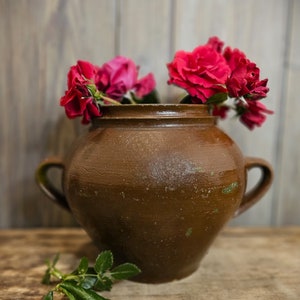 以下が含まれることがあります： 2つの取っ手が付いた茶色の陶器の壺に赤いバラが活けられています。壺は木の表面の上に置かれています。