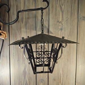 May include: A black metal lantern with a decorative design hangs from a chain attached to a black metal hook. The lantern has a square shape with a pointed top and a cage-like design.