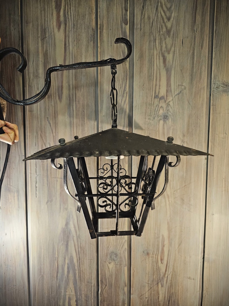 May include: A black metal lantern with a decorative design hangs from a chain attached to a black metal hook. The lantern has a square shape with a pointed top and a cage-like design.