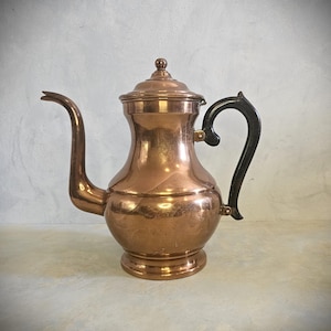 Op de afbeelding: Een koperkleurige koffiepot met een gebogen tuit en een zwarte handgreep. De pot heeft een deksel met een kleine knop bovenop. De pot is gemaakt van glanzend koper en heeft een klassiek ontwerp.