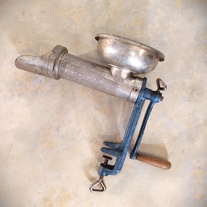 May include: A vintage food mill featuring a silver-toned metal hopper and a perforated cylindrical body. The device includes a blue cast iron clamp mechanism for attachment to a surface, a hand-crank handle with a wooden grip, and a metal screw.