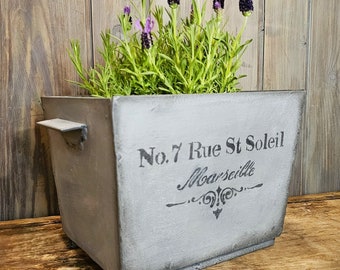 Op de afbeelding: Een grijze metalen plantenbak met een verweerde afwerking en een handvat. De plantenbak heeft de tekst "No. 7 Rue St Soleil Marseille" op de voorkant gedrukt. De plantenbak is gevuld met lavendelplanten.
