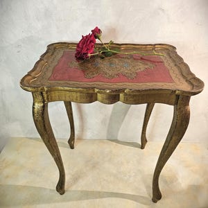 May include: A small, antique, square table with ornate gold trim and legs. The tabletop features a red center with a gold and blue design. A red rose rests on the table's surface. The table is approximately 24 inches long, 24 inches wide, and 20 inches tall.