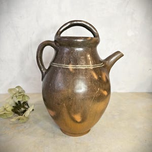 May include: A brown ceramic pitcher with a handle and spout. The pitcher has a rounded body and a loop handle at the top. A smaller handle is on the side. The pitcher has a light-colored band around the middle.
