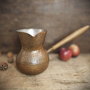 May include: A hammered copper coffee pot with a wooden handle. The pot has a rounded body and a wide, flared spout. The handle is attached to the side of the pot.