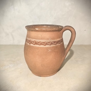 May include: A small, handmade terracotta mug with a handle. The mug features a decorative zigzag pattern around the middle and a slightly darker rim. The overall color is a warm, earthy tone.